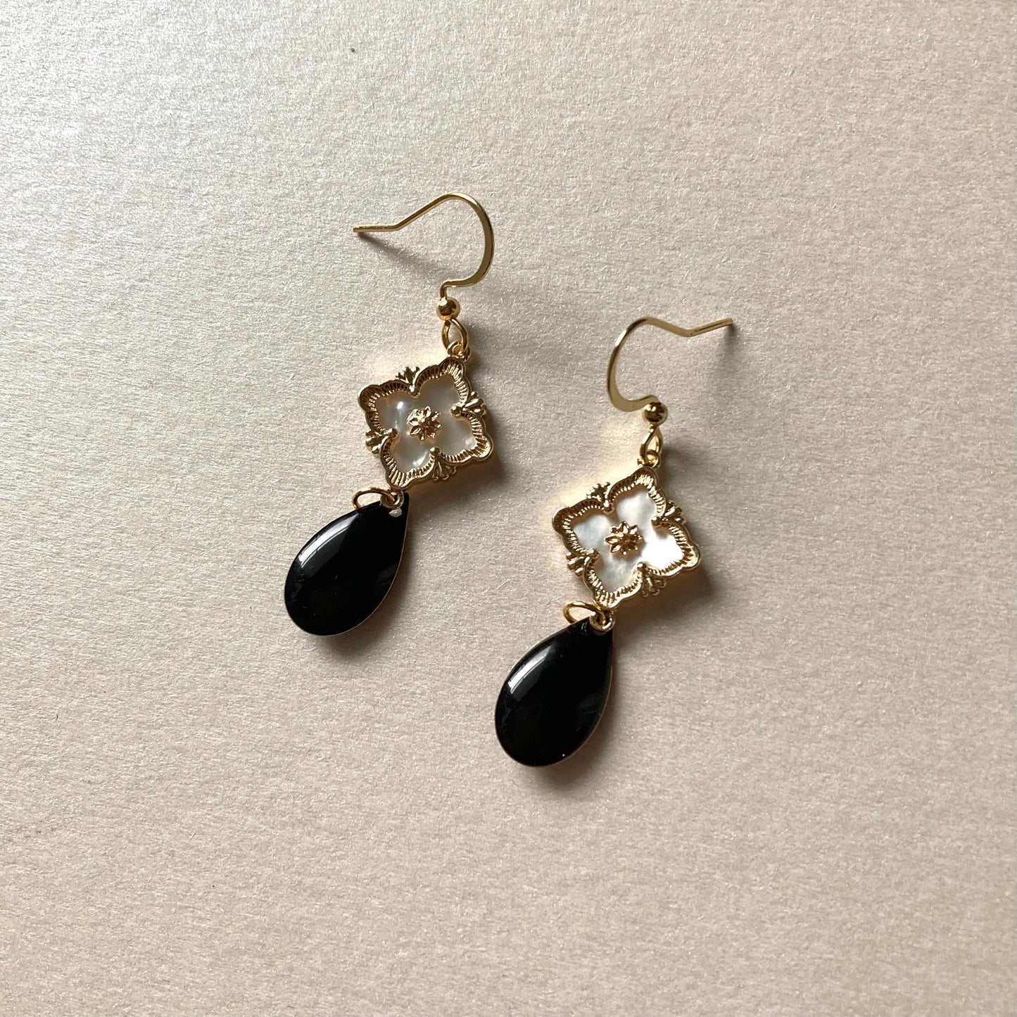 Goldfarbene Ohrringe mit filigranem Blumenmotiv und schwarzem, tropfenförmigem Anhänger auf hellem Untergrund.