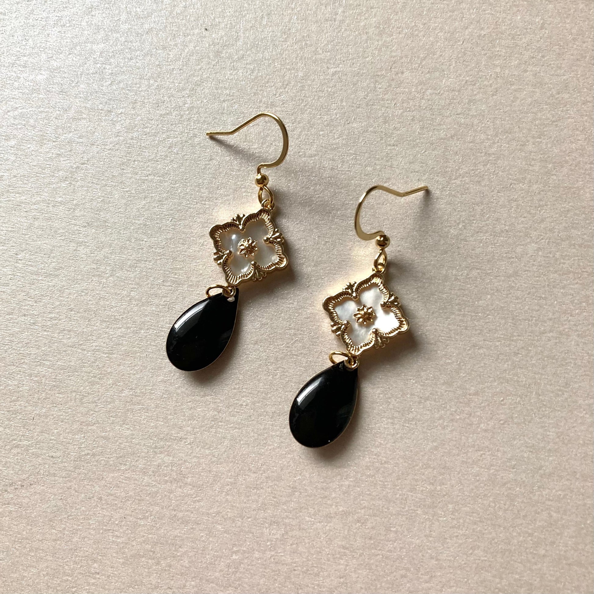 Goldfarbene Ohrringe mit filigranem Blumenmotiv und schwarzem, tropfenförmigem Anhänger auf hellem Untergrund.