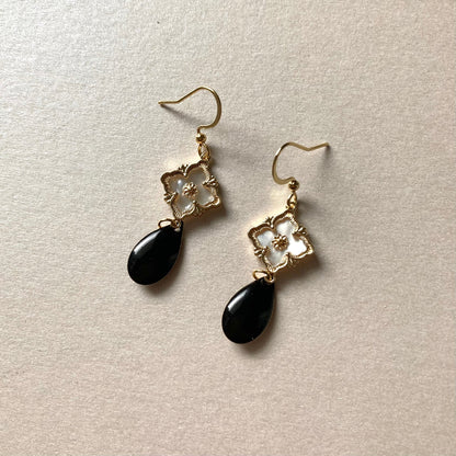Goldfarbene Ohrringe mit filigranem Blumenmotiv und schwarzem, tropfenförmigem Anhänger auf hellem Untergrund.