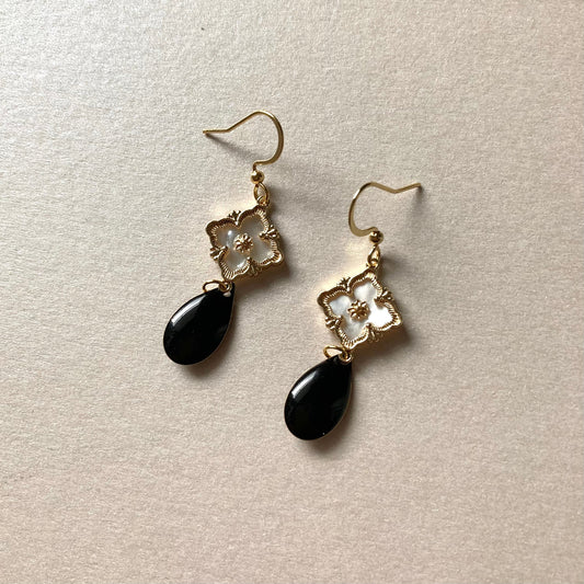 Goldfarbene Ohrringe mit filigranem Blumenmotiv und schwarzem, tropfenförmigem Anhänger auf hellem Untergrund.