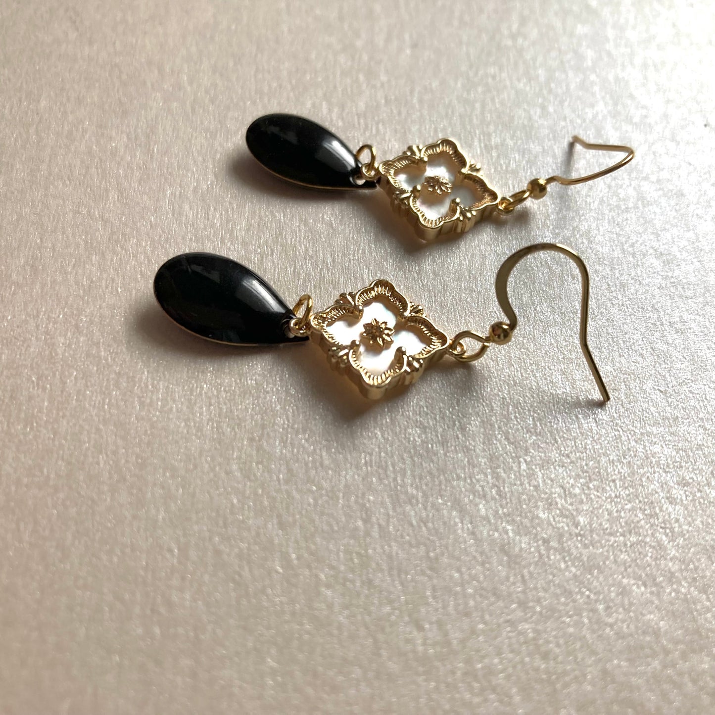 Goldfarbene Ohrringe mit filigranem Blumenmotiv und schwarzem, tropfenförmigem Anhänger auf hellem Untergrund.