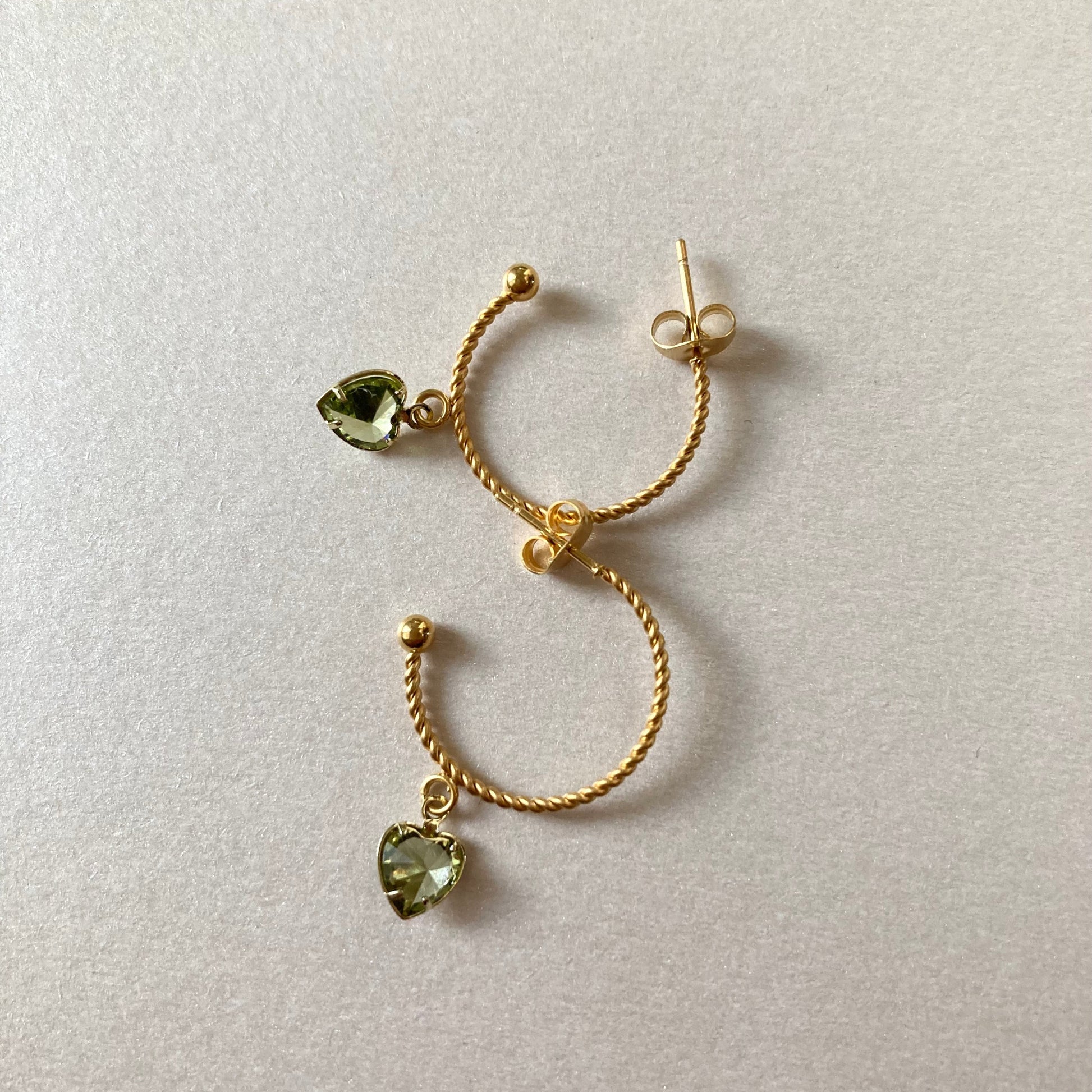 Goldfarbene Creolen mit gedrehter Struktur und herzförmigen, hellgrünen Anhängern liegen auf hellem Untergrund.