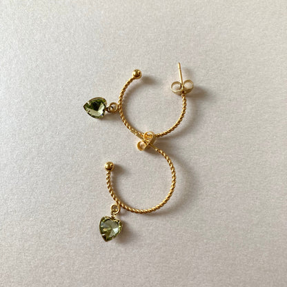 Goldfarbene Creolen mit gedrehter Struktur und herzförmigen, hellgrünen Anhängern liegen auf hellem Untergrund.