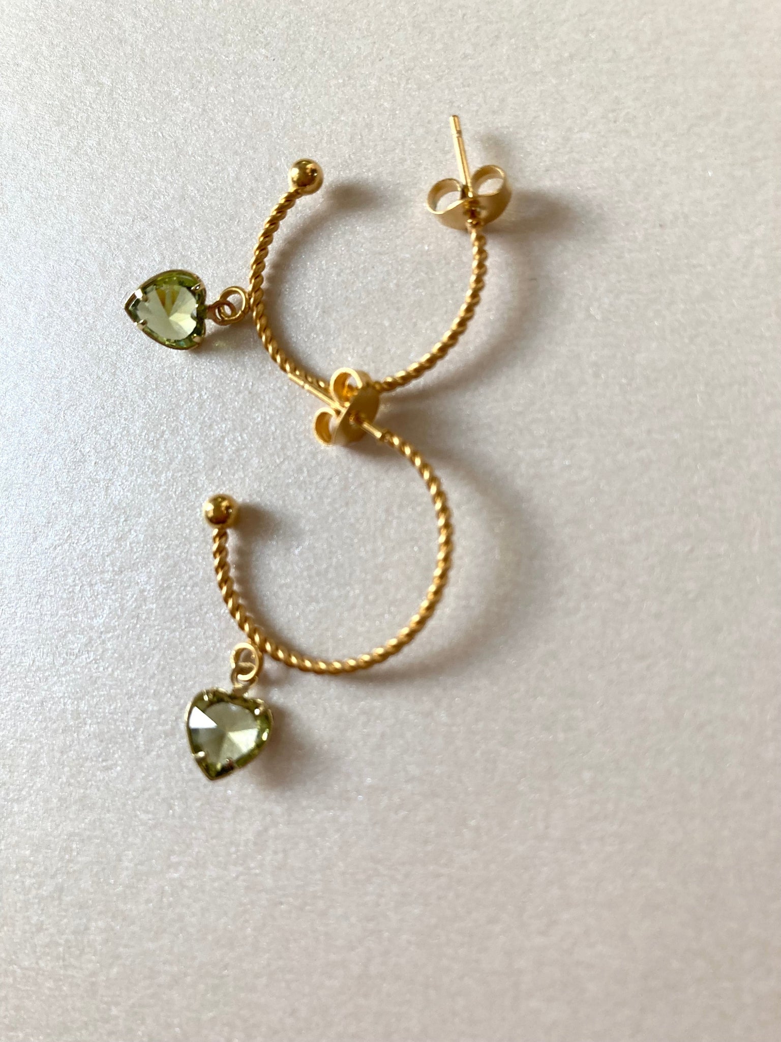 Goldfarbene Creolen mit gedrehter Struktur und herzförmigen, hellgrünen Anhängern liegen auf hellem Untergrund.