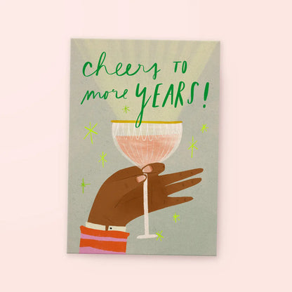 Hand mit dunkler Haut hält ein Sektglas, darüber Schrift 'cheers to more YEARS!' in grüner Schrift