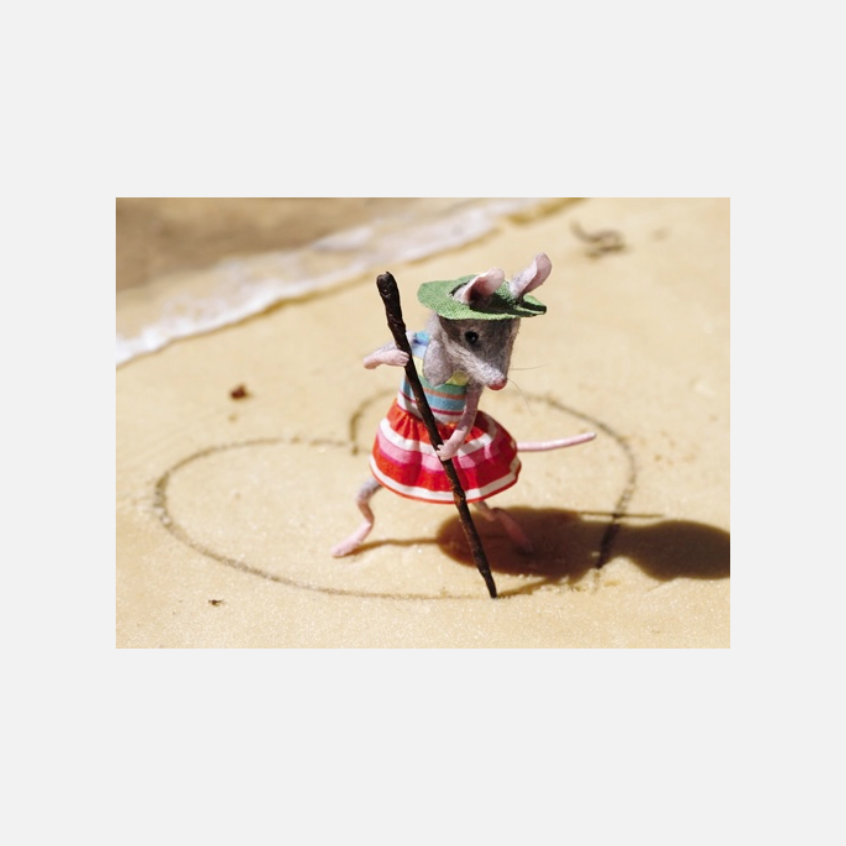 Mausfigur mit grünem Hut, gestreiftem Oberteil und rotem Rock hält einen Stock und steht auf Sand und malt ein Herz 