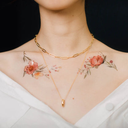 Schulterbereich einer Frau mit floralen Tattoos in Rot- und Orangetönen auf beiden Seiten, zwei goldene Halsketten, weiße Kleidung