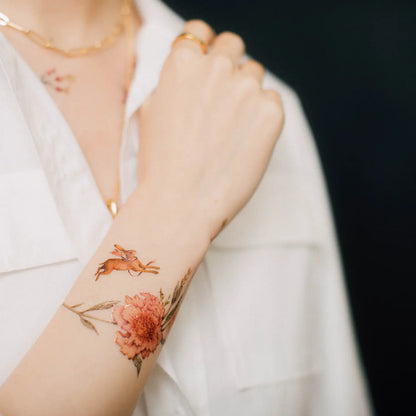 Unterarm mit temporärem Tattoo eines fliegenden Hasen und einer großen Blume, Person trägt weiße Bluse und goldene Kette