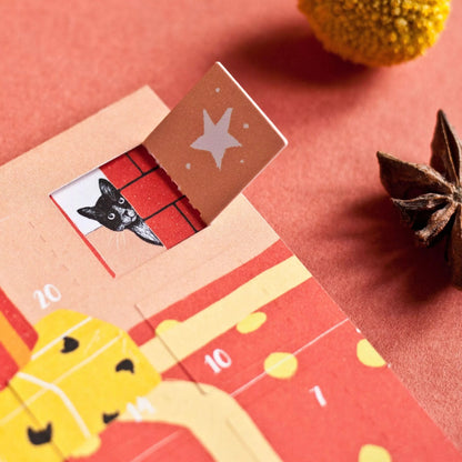 Adventskalender mit schwarzer Katze, die auf große rot-gelbe Geschenke mit Punkten springt, umgeben von Tannenzapfen und Beeren auf rotem Untergrund.
