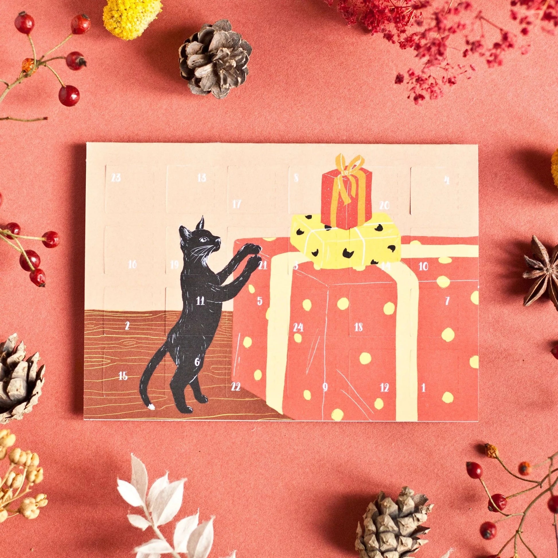 Adventskalender mit schwarzer Katze, die auf große rot-gelbe Geschenke mit Punkten springt, umgeben von Tannenzapfen und Beeren auf rotem Untergrund.