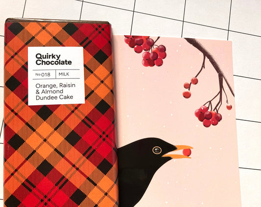 Quirky Chocolate Nr. 018 Vollmilchschokolade „Dundee Cake“ mit Orange, Rosinen und Mandeln in orange, rot und schwarz karierter Verpackung neben einer illustrierten Grußkarte mit einem schwarzen Vogel mit roten Beeren