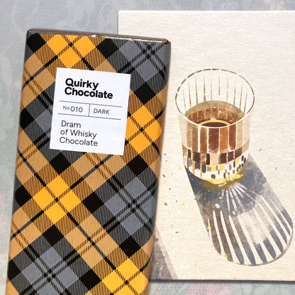 Quirky Chocolate Nr. 010, dunkler Schokoladenriegel „Dram of Whisky“ in gelb-grau-schwarz karierter Verpackung neben einem Glas Whisky auf einem hölzernen Servierbrett mit Schatten.