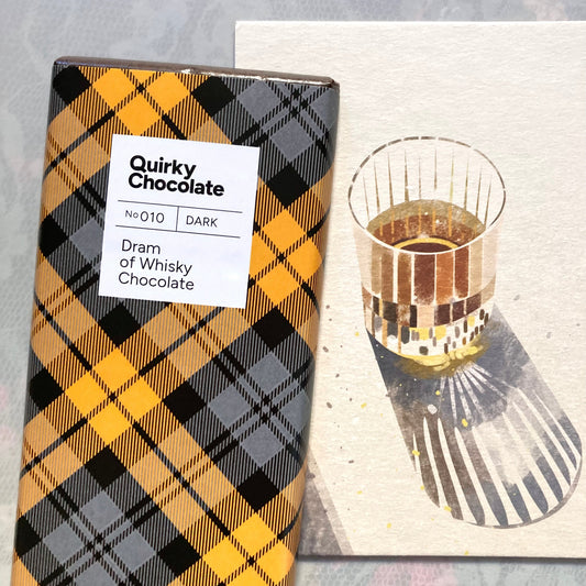 Quirky Chocolate Nr. 010, dunkler Schokoladenriegel „Dram of Whisky“ in gelb-grau-schwarz karierter Verpackung neben einem Glas Whisky auf einem hölzernen Servierbrett mit Schatten.