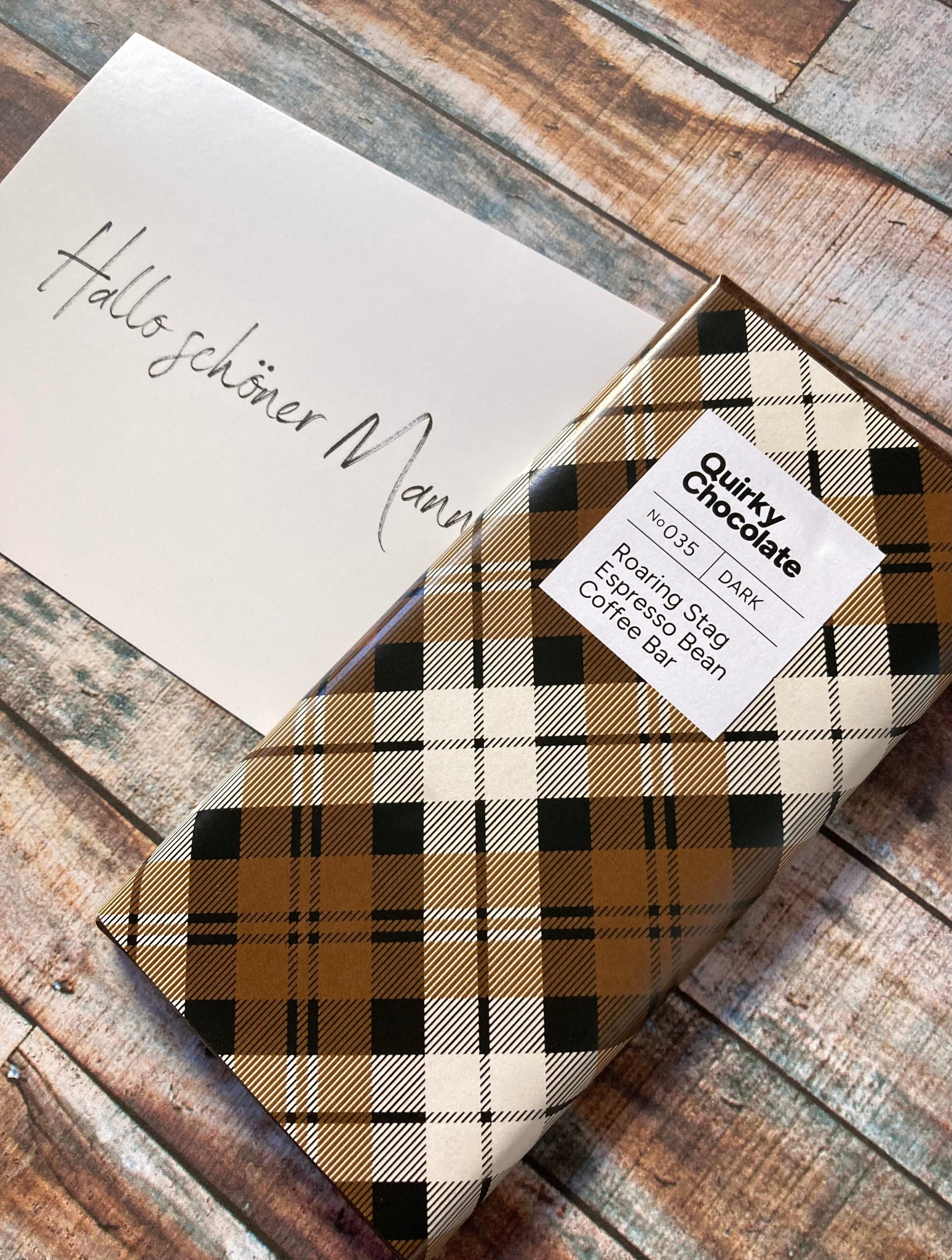 Quirky Chocolate Nr. 035, Kaffee Schokolade aus gerösteten Espressobohnen und aus dunkler Schokolade in braun-schwarz karierter Verpackung auf verwitterten Holzbrettern mit handgeschriebener Grußkarte „Hallo schöner Mann“.