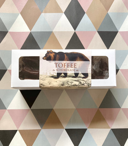 Weiße Schachtel mit durchsichtigen Fenstern, darin mehrere dunkle Toffee-Pralinen, Etikett mit braunem Bären und Schrift 'TOFFEE SCHOKOLADE'.