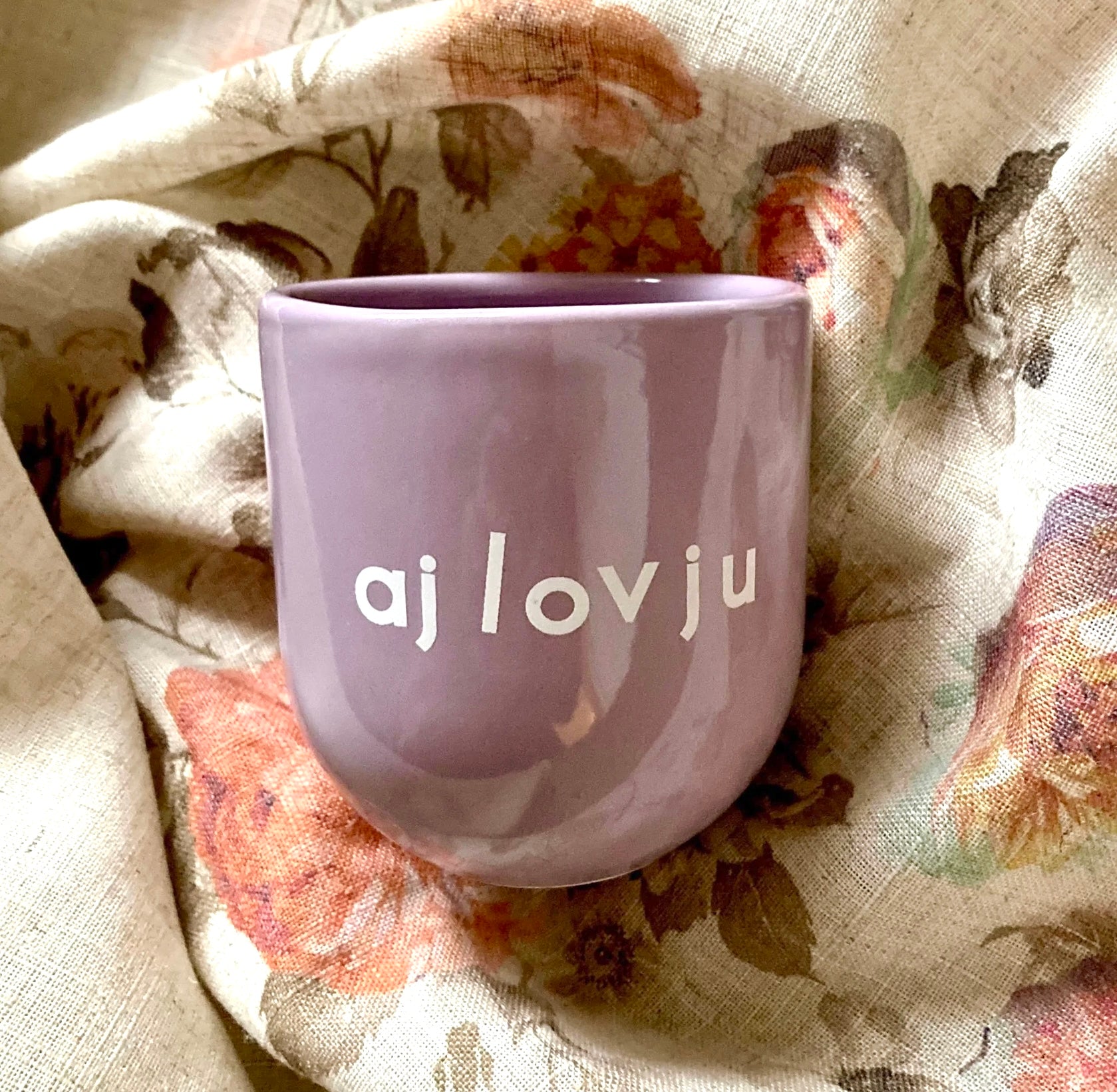 Ein lilafarbener Keramikbecher mit weißer Aufschrift „aj lov ju“ auf geblümter Decke.