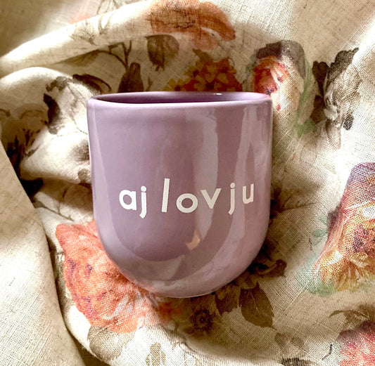 Ein lilafarbener Keramikbecher mit weißer Aufschrift „aj lov ju“ auf geblümter Decke.