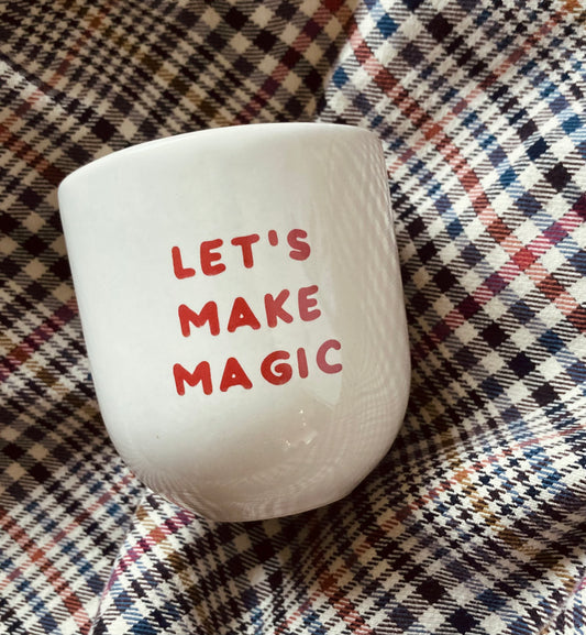 Ein weißer Keramikbecher mit der Aufschrift ‚LET’S MAKE MAGIC‘ in roter Schrift, die auf einem karierten Stoff oder einer Decke mit einem mehrfarbigen Muster aus Streifen und Karos in den Farben Rot, Blau, Schwarz, Weiß und Braun steht.