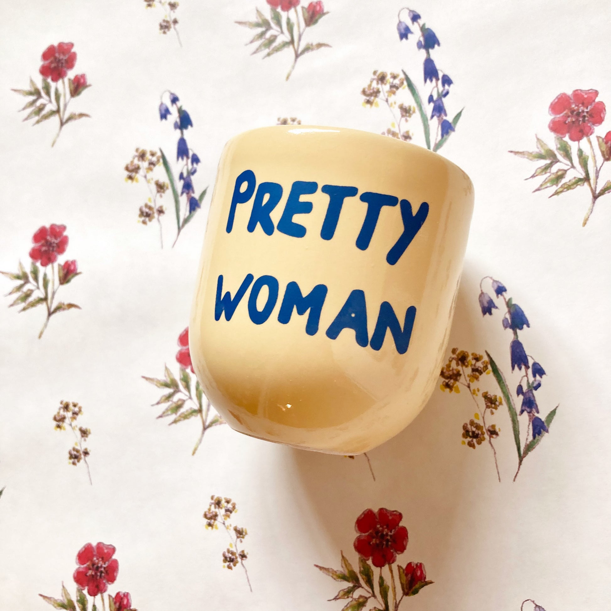 „Ein gelber Keramikbecher mit der blauen Aufschrift ‚PRETTY WOMAN‘ auf einer weißen Oberfläche mit zartem Blumenmuster aus Papier