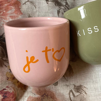 Ein rosa Keramikbecher mit orangefarbener Aufschrift „je taime“ auf geblümter Decke.
