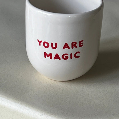 Ein weißer Keramikbecher mit der Aufschrift ‚LET’S MAKE MAGIC‘ in roter Schrift.