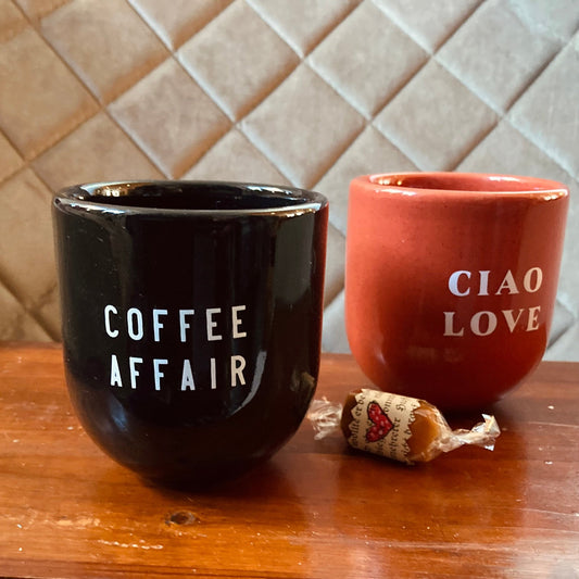 Zwei Keramikbecher stehen auf einer Holzoberfläche vor einer beigefarbenen Fliesenrückwand mit Rautenmuster. der linke becher ist glänzend schwarz mit der weißen Aufschrift „COFFEE AFFAIR“, der rechte Becher ist korallen- oder orangerot mit der weißen Aufschrift „CIAO LOVE“. Zwischen den Tassen auf der Holztheke steht ein eingewickeltes Bonbon.