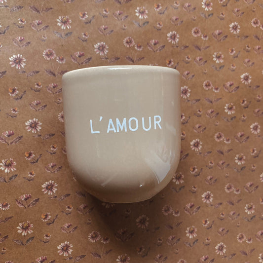 Ein hellbrauner Keramikbecher mit weißer Aufschrift „L’AMOUR“ auf braunem Papier im Vintage-Blumenmuster mit kleinen rosa und weißen, gänseblümchenartigen Blüten.