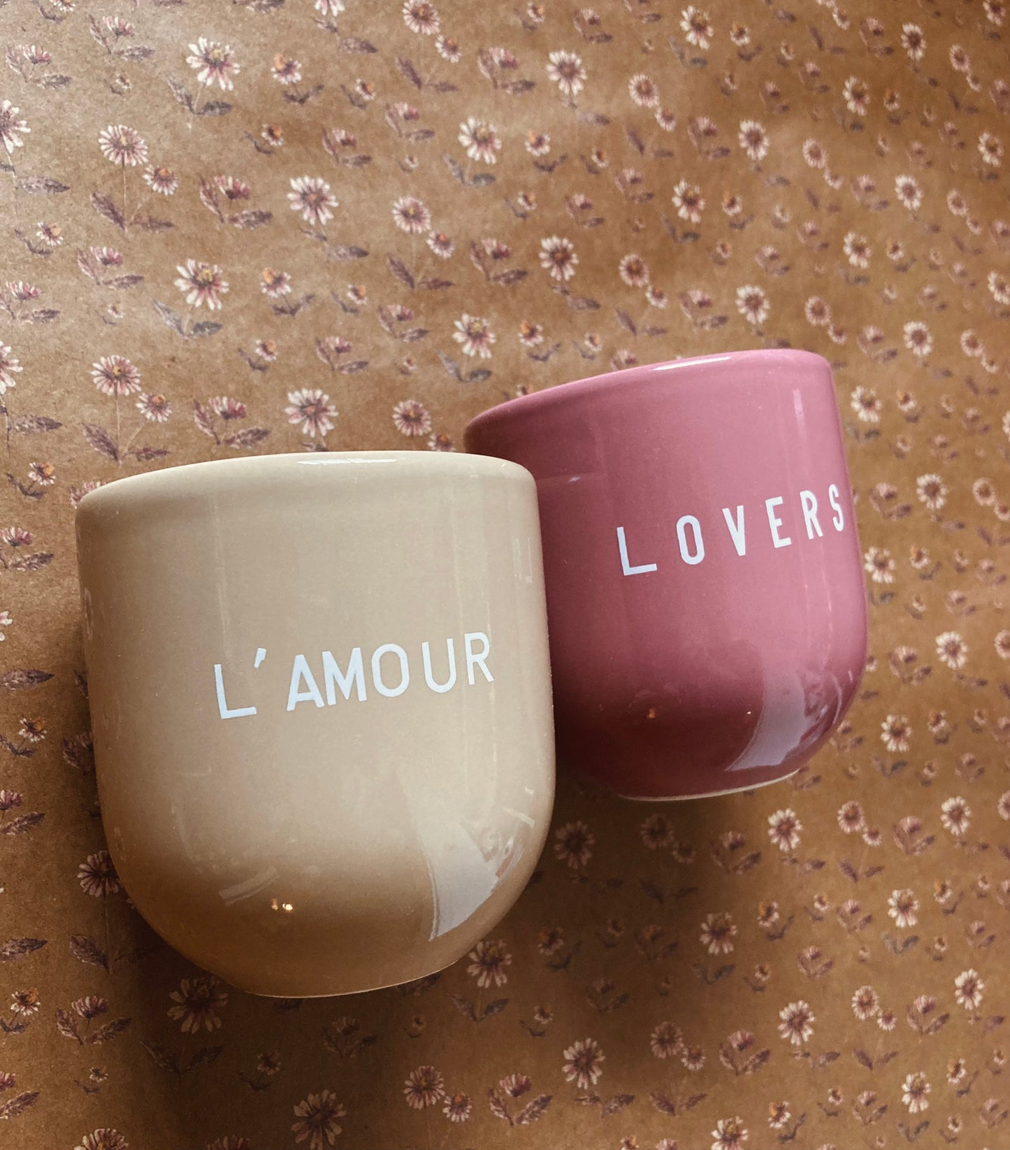 Ein hellbrauner Keramikbecher mit weißer Aufschrift „L’AMOUR“ und ein altrosa Becher mit weißer Aufschrift ‚Lovers’ auf braunem Papier im Vintage-Blumenmuster mit kleinen rosa und weißen, gänseblümchenartigen Blüten.