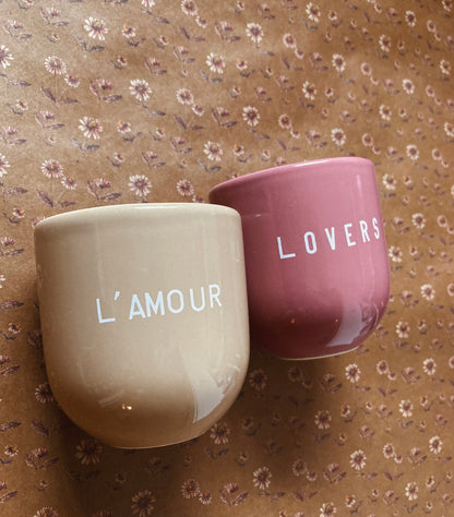Ein hellbrauner Keramikbecher mit weißer Aufschrift „L’AMOUR“ und ein altrosa Becher mit weißer Aufschrift ‚Lovers’ auf braunem Papier im Vintage-Blumenmuster mit kleinen rosa und weißen, gänseblümchenartigen Blüten.