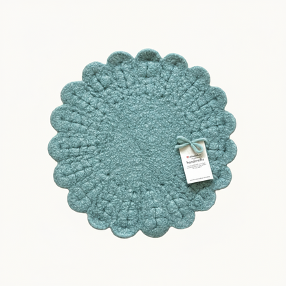 Eine handgehäkelte Sitzunterlage in zartem Salbei mit romantischen Blütensaum. Das zentrale Häkelmuster in Kreisform bildet einen harmonischen Kontrast zum verspielten Rand. 