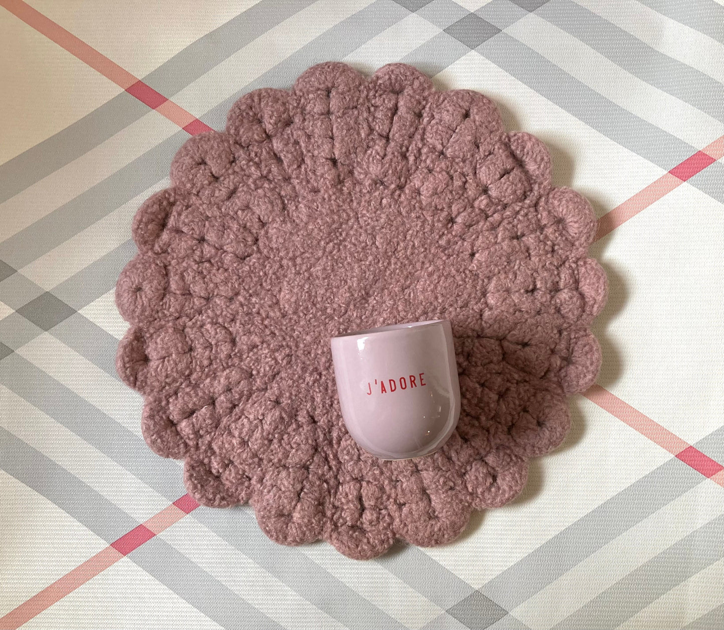 Ein lilafarbener Keramikbecher mit roter Aufschrift „J‘ADORE“ auf gehäkelter Sitzauflage in Rosa
