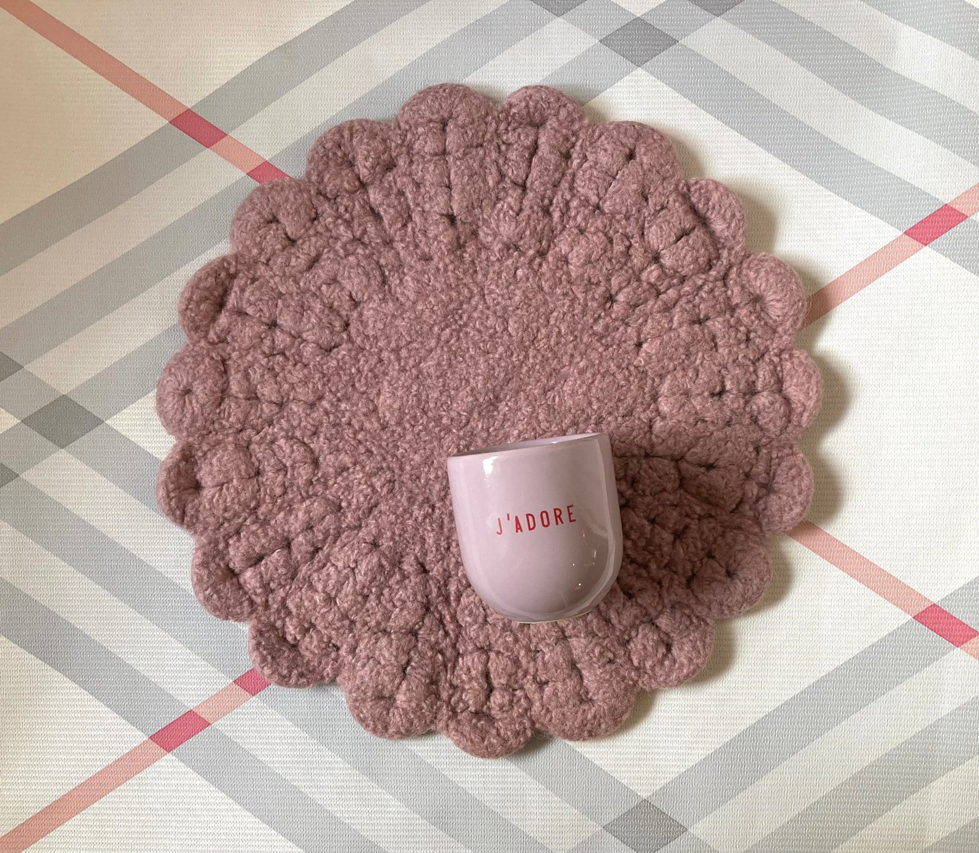 Ein lilafarbener Keramikbecher mit roter Aufschrift „J‘ADORE“ auf gehäkelter Sitzauflage in Rosa