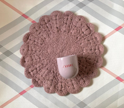 Ein lilafarbener Keramikbecher mit roter Aufschrift „J‘ADORE“ auf gehäkelter Sitzauflage in Rosa