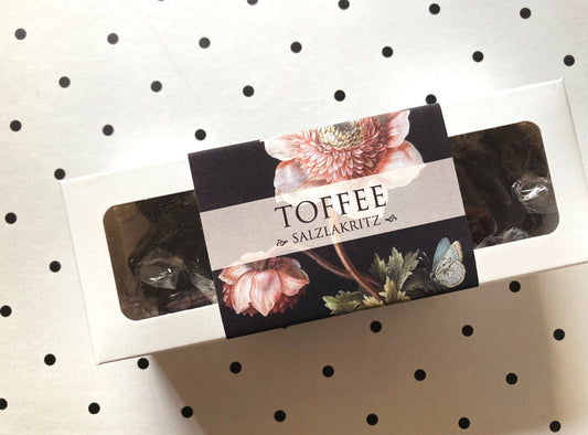 Weiße rechteckige Verpackung mit durchsichtiger Folie, schwarzem Etikett mit Blumenmotiv und Schriftzug 'TOFFEE mit SALZLAKRITZ'.