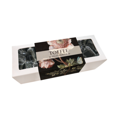 Weiße rechteckige Verpackung mit durchsichtiger Folie, schwarzem Etikett mit Blumenmotiv und Schriftzug 'TOFFEE mit SALZLAKRITZ'.
