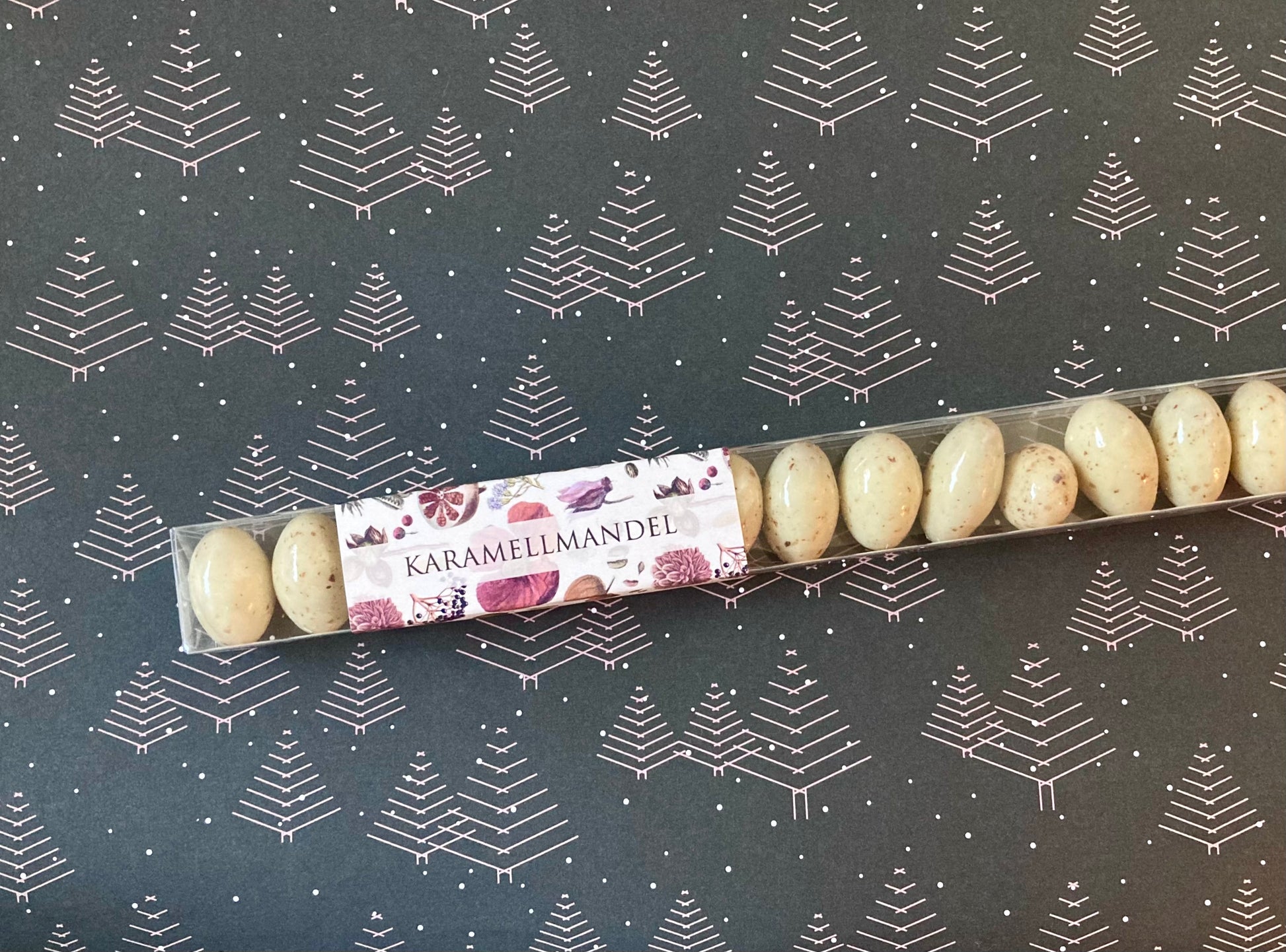 Lange, transparente Verpackung mit hellbraunen Karamellmandeln auf dunkelgrauem Papier mit weißem Tannenbaum-Muster.