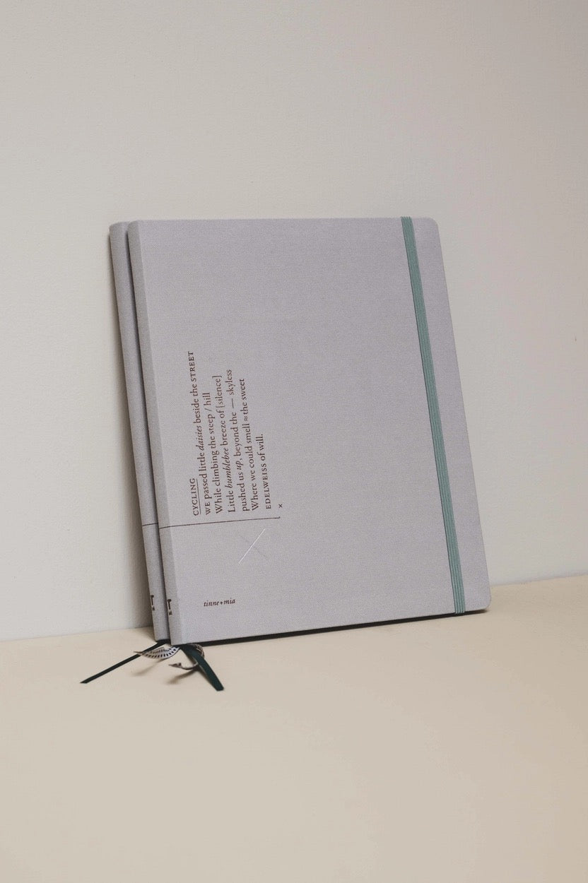 Zwei übereinanderliegende hellgraue Planerr mit graugrünem Gummiband und drei Lesebändchen in Schwarz und Weiß. 