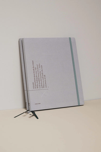 Zwei übereinanderliegende hellgraue Planerr mit graugrünem Gummiband und drei Lesebändchen in Schwarz und Weiß. 