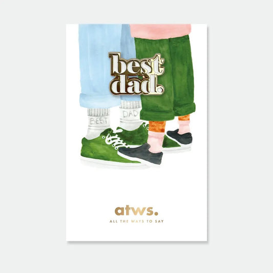 Ein goldener Emaille-Pin mit dem Schriftzug „best dad“ auf einer illustrierten Karte. Die Illustration zeigt die Beine und Füße eines Erwachsenen und eines Kindes, die einander gegenüberstehen; der Vater trägt grüne Sneaker und Socken mit der Aufschrift „Best Dad“.