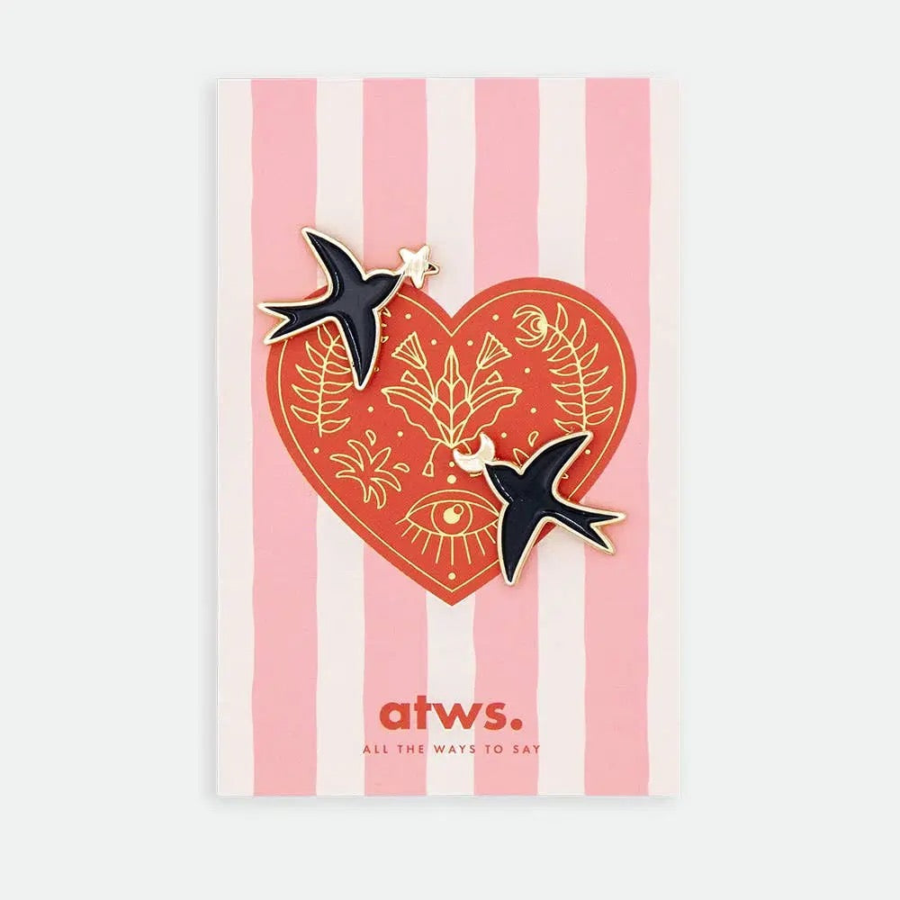 Produktgrafik eines Pin-Sets von „All The Ways To Say“. Zwei schwarze Schwalben-Pins mit Goldrand sind auf einer Karte befestigt. Die Karte zeigt ein rotes Herz mit goldenen floralen Illustrationen und einem Auge auf einem Hintergrund aus zartrosa Längsstreifen.