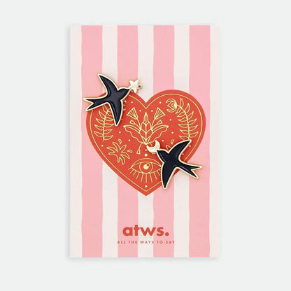 Produktgrafik eines Pin-Sets von „All The Ways To Say“. Zwei schwarze Schwalben-Pins mit Goldrand sind auf einer Karte befestigt. Die Karte zeigt ein rotes Herz mit goldenen floralen Illustrationen und einem Auge auf einem Hintergrund aus zartrosa Längsstreifen.