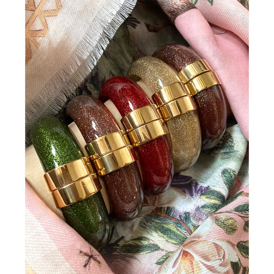 Fünf funkelnde Glitzer-Armreife in den Farben Smaragdgrün, Cognacbraun, Rubinrot, Champagner-Gold und Kupferbraun, nebeneinander arrangiert auf einem hellen Displaykissen. Die Armreife aus transparentem Kunstharz verfügen jeweils über eine markante, polierte Klappschließe in Gold-Optik und liegen dekorativ eingebettet zwischen einem zartrosa gemusterten Seidentuch und einem beigen Textilgewebe.