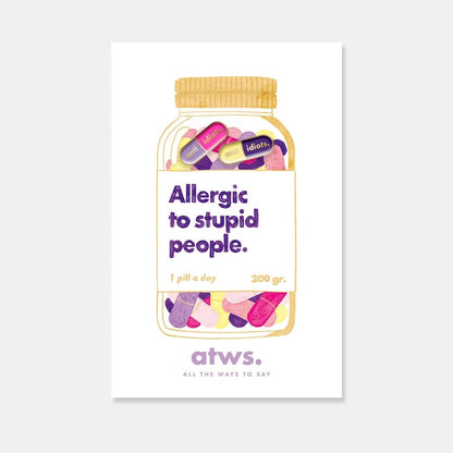 Zwei Emaille-Pins in Pillenform (lila/pink und gelb/dunkelblau) mit der Aufschrift „anti idiots“ auf einer Trägerkarte, die ein Glas mit der Aufschrift „Allergic to stupid people“ zeigt.