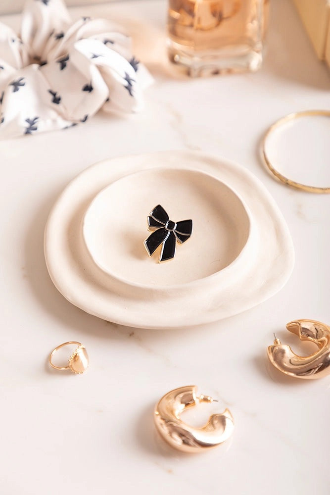 Eleganter Emaille-Pin in Form einer großen schwarzen Schleife mit feiner goldener Umrandung auf einem Teller.