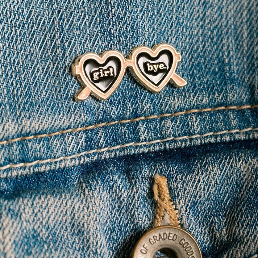 Emaille-Pin in Form einer herzförmigen Sonnenbrille, auf deren Gläsern „girl“ und „bye“ steht, präsentiert auf einer Jeansjacke