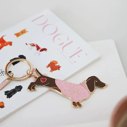 Nahaufnahme des Dackel-Schlüsselanhängers von All the ways to Say auf einem Notizbuch. Der Anhänger zeigt einen Dackel im rosa Sweater mit Glitzer-Effekt und einem roten Herz-Detail am Halsband vor hellem Hintergrund.