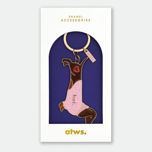 All the ways to Say Emaille-Schlüsselanhänger „Hot Dog“: Ein brauner Dackel mit einem rosa Glitzer-Pullover und der Aufschrift „hot.“, präsentiert in einer weißen Geschenkverpackung mit goldenem Logo.