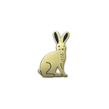 Detailaufnahme eines goldfarbenen Emaille-Pins in Form eines sitzenden Feldhasen mit schwarzen Akzenten an den Ohren und minimalistischen grafischen Details