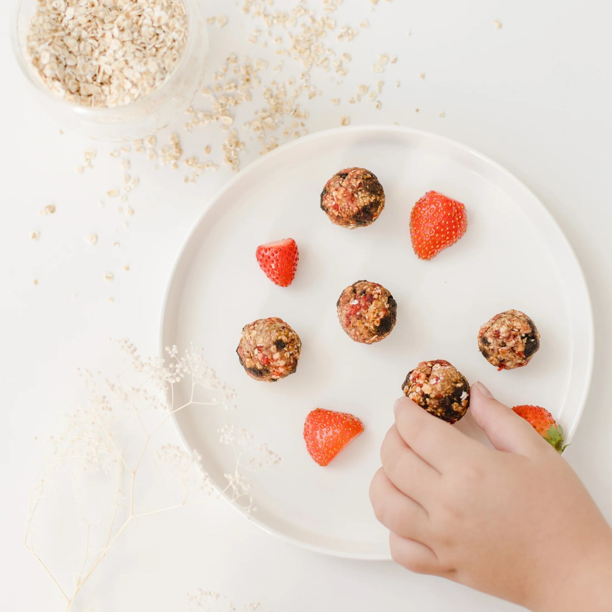 Hand greift nach einem Energiebällchen auf weißem Teller mit mehreren Erdbeerstücken und Energiebällchen.