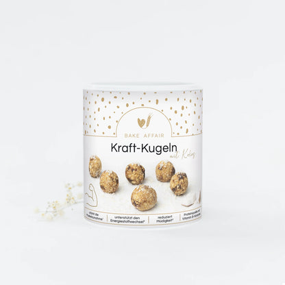 Runde Kraft-Kugeln mit Kokos auf weißem Verpackungsbecher von Bake Affair, umgeben von goldfarbenen Punkten.